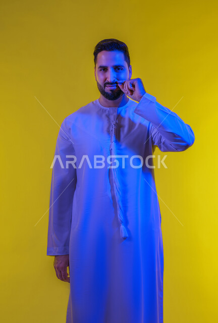 إيماءات الالتزام والوفاء بالوعود، حركات وتعابير شبابيه، بورتريه لرجل عربي خليجي اماراتى يرتدي الكندورة ينظر الى الكاميرا ويمسك شاربه، مفهوم الرجولة والقوة، التهديد والتوعد لشيء ما، الطول الكامل للجسم، خلفية صفراء .