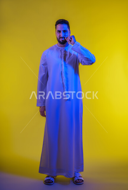 مفهوم الرجولة والقوة، التهديد والتوعد لشيء ما، الاعجاب بنمو الشارب، بورتريه لرجل عربي خليجي اماراتى يرتدي الكندورة ينظر الى الكاميرا ويمسك شاربه، إيماءات الالتزام والوفاء بالوعود، حركات وتعابير شبابيه، الطول الكامل للجسم، خلفية صفراء .