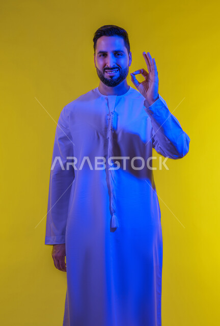 رفع اليد بعلامات الجودة والقبول، الامتياز والعلامة الكاملة، التعبير عن الإعجاب والتقدير، الشعور بالرضا والاستحسان، بورتريه لرجل عربي خليجي اماراتي يرتدي الكندورة ينظر إلى الكاميرا بإيماءات السعادة والسرور، الجسم بالطول الكامل، خلفية صفراء .