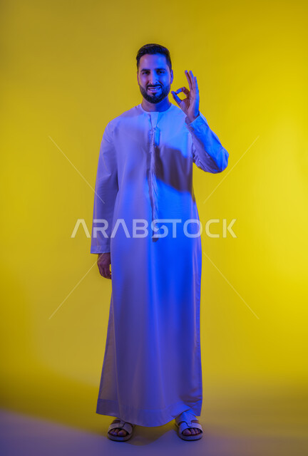 التعبير عن الإعجاب والتقدير، الامتياز والعلامة الكاملة، رفع اليد بعلامات الجودة والقبول، الشعور بالرضا والاستحسان، بورتريه لرجل عربي خليجي اماراتي يرتدي الكندورة ينظر إلى الكاميرا بإيماءات السعادة والسرور، الجسم بالطول الكامل، خلفية صفراء .