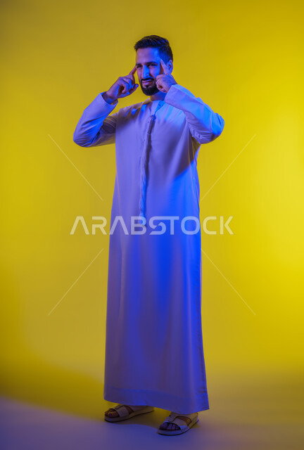 الاحساس بالشقيقية والصداع النصفي, الاجهاد والضغط النفسي, الإرهاق وفقدان التركيز, الشعور بالصداع وألم الرأس, بورتريه لرجل عربي خليجي اماراتي يرتدي الكندورة يضع يديه على رأسه ويغمض عينيه بإيماءات الألم الشديد, صورة الجسم بالطول الكامل, خلفية صفراء .