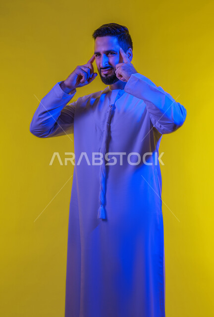 الإرهاق وفقدان التركيز, الشعور بالصداع وألم الرأس, الاحساس بالشقيقية والصداع النصفي, الاجهاد والضغط النفسي, بورتريه لرجل عربي خليجي اماراتي يرتدي الكندورة يضع يديه على رأسه ويغمض عينيه بإيماءات الألم الشديد, صورة الجسم بالطول الكامل, خلفية صفراء .