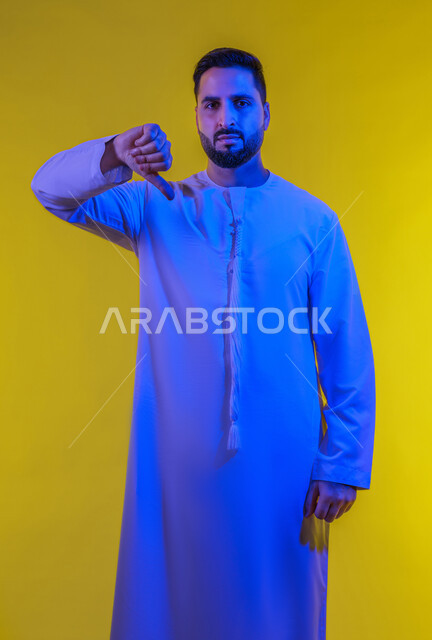 إيماءات عدم الاعجاب والتقييم السلبي ، الشعور بالانزعاج من شيء ما، حركات يدين تدل على الرفض وعدم القبول والفشل، بورتريه لرجل عربي خليجي امارتي يرتدي الكندورة يشير بإصبعه الابهام الى الأسفل ، صورة الجسم بالطول الكامل, خلفية صفراء .