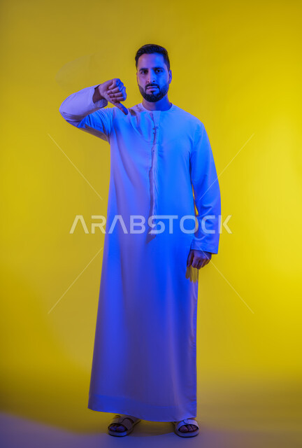 الشعور بالانزعاج من شيء ما، حركات يدين تدل على الرفض وعدم القبول والفشل، بورتريه لرجل عربي خليجي امارتي يرتدي الكندورة يشير بإصبعه الابهام الى الأسفل بإيماءات عدم الاعجاب والتقييم السلبي، صورة الجسم بالطول الكامل, خلفية صفراء .