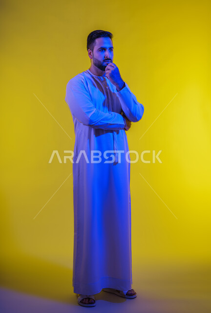 الشعور بالحيرة وعدم التأكد, إيماءات التأمل والتمعن والتركيز, بورتريه لرجل عربي خليجي اماراتي يرتدي يرتدي الثوب الكندورة يقف ويضع يده تحت ذقنه، النظر الى مكان ما بتعابير التفكير، التخطيط لحل مشكلة ما،الجسم بالطول الكامل، خلفية صفراء .