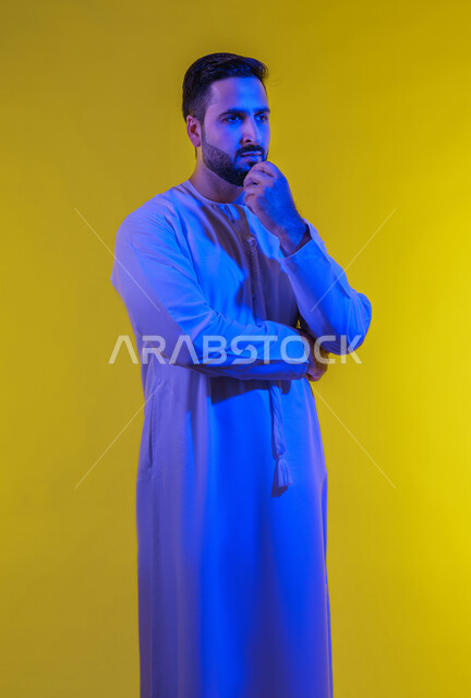 إيماءات التأمل والتمعن والتركيز, بورتريه لرجل عربي خليجي اماراتي يرتدي يرتدي الثوب الكندورة يقف ويضع يده تحت ذقنه، النظر الى مكان ما بتعابير التفكير، التخطيط لحل مشكلة ما، الشعور بالحيرة وعدم التأكد, الجسم بالطول الكامل، خلفية صفراء .