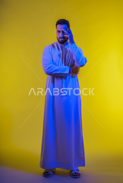 Fatigue, exhaustion and psychological pressure, gestures of thinking and anxiety, portrait of a young Arab Gulf Emirati man wearing a kandura, putting his hand on his face, feeling a bad state of tingling and pain, full-length body image, yellow background.
