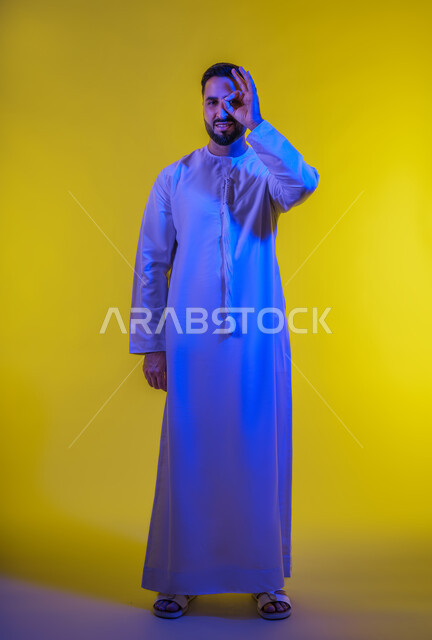 رفع اليد بعلامات الجودة والقبول، الامتياز والعلامة الكاملة، التعبير عن الإعجاب والتقدير،الشعور بالرضا والاستحسان، بورتريه لرجل عربي خليجي اماراتي يرتدي الكندورة ينظر إلى الكاميرا بإيماءات السعادة والسرور، الجسم بالطول الكامل، خلفية صفراء .