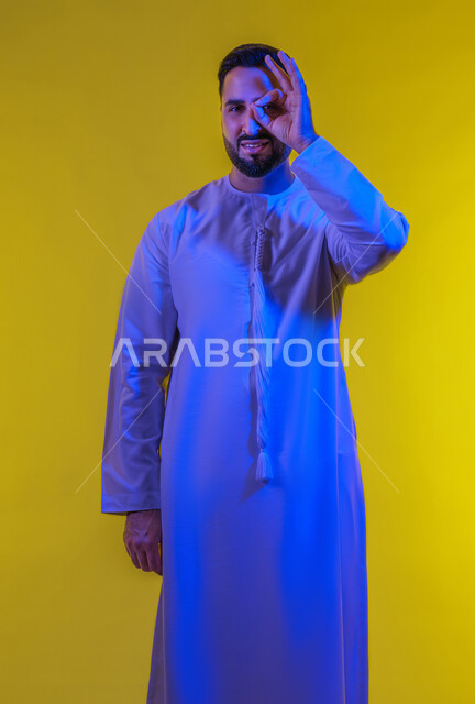 الامتياز والعلامة الكاملة، التعبير عن الإعجاب والتقدير، رفع اليد بعلامات الجودة والقبول، الشعور بالرضا والاستحسان، بورتريه لرجل عربي خليجي اماراتي يرتدي الكندورة ينظر إلى الكاميرا بإيماءات السعادة والسرور، الجسم بالطول الكامل، خلفية صفراء .