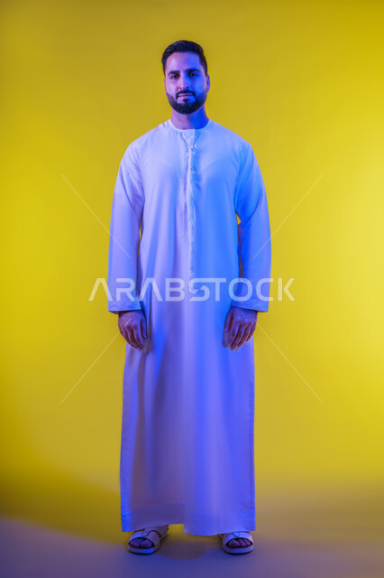 التقاط الصور الشخصية للوثائق الرسمية, مفهوم الاناقة والرجولة, بورتريه لرجل عربي خليجي إماراتي مبتسم يرتدي الكندورة يقف باستقامة بتعابير البشاشة والسرور, الاهتمام بالمظهر الخارجي، النظر الى الكاميرا بايماءات الثقة بالنفس, خلفية صفراء .