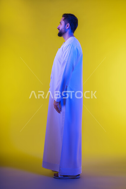 تعابير الجدية والحزم,الاهتمام بالمظهر الخارجي، بورتريه من الجانب لرجل عربي خليجي إماراتي يرتدي الكندورة الاماراتية ينظر الى شيء ما, الوقوف باستقامة وثبات والنظر الى الشمال بايماءات الثقة بالنفس, مفهوم الأناقة والرجولة والقوة خلفية صفراء.
