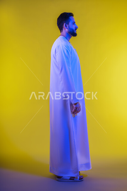 مفهوم الأناقة والرجولة والقوة, بورتريه من الجانب لرجل عربي خليجي إماراتي يرتدي الكندورة الاماراتية ينظر الى شيء ما, الوقوف باستقامة وثبات والنظر الى اليمين بايماءات الثقة بالنفس, تعابير الجدية والحزم،الاهتمام بالمظهر الخارجي، خلفية صفراء.