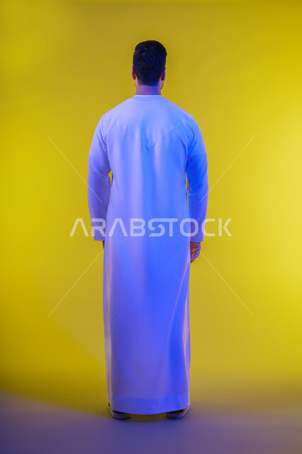الوقوف باستقامة وثقة بالنفس , الاهتمام بالاناقة والمظهر الخارجي, بورتريه من الخلف لرجل عربي خليجي امارتي يرتدي الكندورة , ارتداء الملابس التقليدية، صورة الجسم بالطول الكامل ، خلفية صفراء .