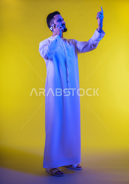 اجراء مكالمة هاتفية من خلال الجوال، بورتريه لرجل عربي خليجي إماراتي يرتدي الكندورة الإماراتية يشير باصبع السبابة لاعلى ، استخدام الهاتف المحمول في الاتصال والتواصل مع الأفراد، دردشة ومحادثات وتعارف،صورة الجسم بالطول الكامل ، خلفية صفراء .