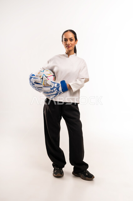 Training and warm-ups before the match, professional team sports, practicing the hobby of playing football, portrait of a young Saudi Arabian Gulf Arab woman wearing goalkeeper gloves, holding a football in her hand with expressions and gestures of pleasure and happiness, full body photo, white background