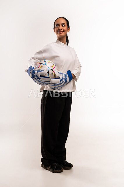 Training and warm-ups before the match, professional team sports, practicing the hobby of playing football, portrait of a young Saudi Arabian Gulf Arab woman wearing goalkeeper gloves, holding a football in her hand with expressions and gestures of pleasure and happiness, full body photo, white background