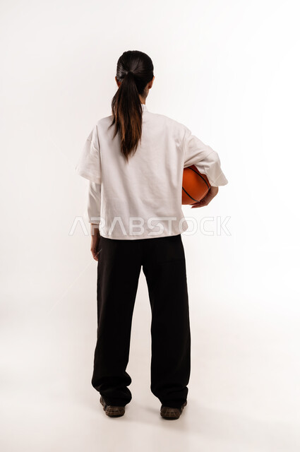 ممارسة هواية كرة السلة, الشغف بالألعاب الرياضية, بورتريه من الخلف لشابة عربية خليجية سعودية ترتدي ملابس رياضية تمسك كره السله بإيماءات التشجيع و الحماس, الفريق السعودي النسائي, صورة للجسم بالطول الكامل, خلفية بيضاء