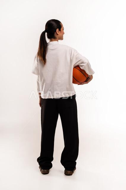 ممارسة هواية كرة السلة, الشغف بالألعاب الرياضية, بورتريه من الخلف لشابة عربية خليجية سعودية ترتدي ملابس رياضية تمسك كره السله بإيماءات التشجيع و الحماس, الفريق السعودي النسائي, صورة للجسم بالطول الكامل, خلفية بيضاء