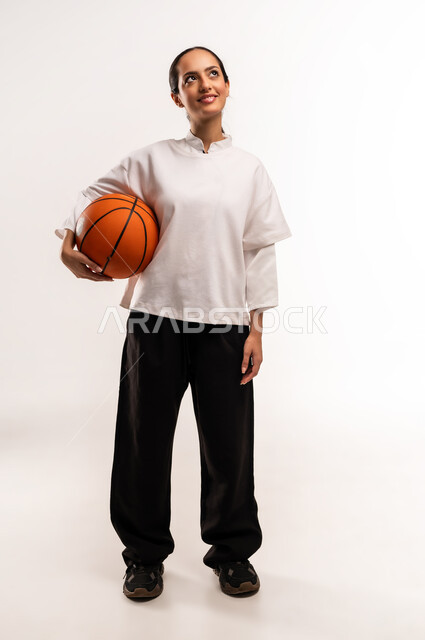 ممارسة هواية كرة السلة, الشغف بالألعاب الرياضية, بورتريه لشابة عربية خليجية سعودية ترتدي ملابس رياضية تمسك كره السله بإيماءات التشجيع و الحماس, الفريق السعودي النسائي, صورة للجسم بالطول الكامل, خلفية بيضاء