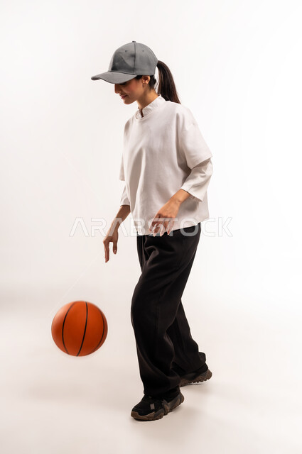 الشغف بالألعاب الرياضية, ممارسة هواية كرة السلة, بورتريه من الجانب لشابة عربية خليجية سعودية ترتدي ملابس رياضية تمسك كره السله بإيماءات التشجيع و الحماس, الفريق السعودي النسائي, صورة للجسم بالطول الكامل, خلفية بيضاء