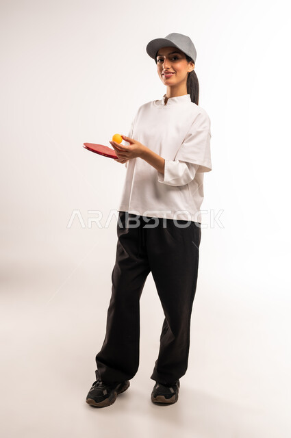 الاستمتاع بالمباريات الودية والبطولات المحلية، ممارسة رياضات وأنشطة بدنية ممتعة، بورتريه لشابة عربية خليجية سعودية ترتدي ملابس رياضية تحمل بيدها مضرب تنس الطاولة، اللعب بسرعة وتركيز عالي، المحافظة على الرشاقة واللياقة الجسدية, الجسم بالطول الكامل، خلفيه بيضاء