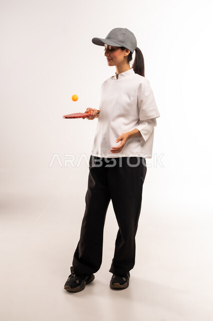 الاستمتاع بالمباريات الودية والبطولات المحلية، ممارسة رياضات وأنشطة بدنية ممتعة، بورتريه لشابة عربية خليجية سعودية ترتدي ملابس رياضية تحمل بيدها مضرب تنس الطاولة، اللعب بسرعة وتركيز عالي، المحافظة على الرشاقة واللياقة الجسدية, الجسم بالطول الكامل، خلفيه بيضاء