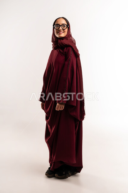 عرض تصاميم موديلات عباءات نسائية معاصرة، بورتريه لعارضة أزياء عربية سعودية خليجية مبتسمة ترتدي الحجاب والعباءة  الحمراء تنظر إلى شيء ما بإيماءات الأنوثة والجمال، مفهوم الموضة والأناقة، متابعة الأزياء الراقية، الجسم بالطول الكامل، خلفية بيضاء