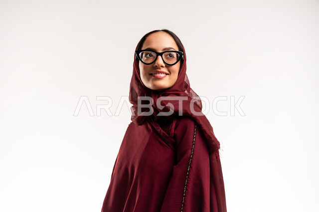 عرض تصاميم موديلات عباءات نسائية معاصرة، بورتريه مقرب لعارضة أزياء عربية سعودية خليجية مبتسمة ترتدي الحجاب والعباءة  الحمراء تنظر إلى شيء ما بإيماءات الأنوثة والجمال، مفهوم الموضة والأناقة، متابعة الأزياء الراقية، خلفية بيضاء