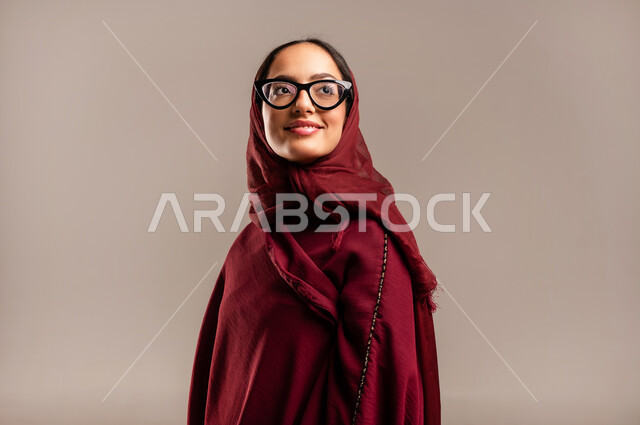 عرض تصاميم موديلات عباءات نسائية معاصرة، بورتريه مقرب لعارضة أزياء عربية سعودية خليجية مبتسمة ترتدي الحجاب والعباءة  الحمراء تنظر إلى الكاميرا بإيماءات تدل على الأنوثة والجمال، مفهوم الموضة والأناقة، متابعة الأزياء الراقية، خلفية بيج