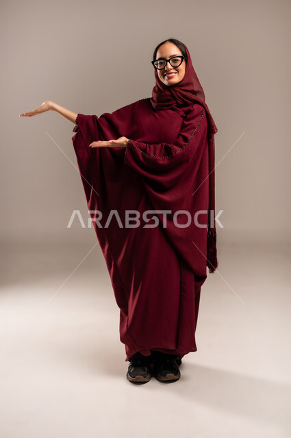 التوضيح والدلالة لمكان محدد, تحديد موقع شيء معين, القاء التحيه والسلام, بورتريه  لامرأة عربية خليجية سعوديه محجبة ترتدي العباءة الحمراء تشير بيديها الى هاذا الاتجاه، بالإشارة إلى عنصر ما, صورة الجسم بالطول الكامل، خلفية بيج