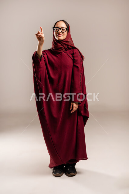 Illustration and indication of a specific place, locating something, greeting and peace, portrait of a veiled Saudi Arabian Gulf woman wearing a red abaya pointing forward with her index finger, a young Saudi woman pointing at an item, full-length body image, beige background