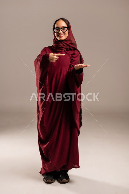 تعابير حمل و إمساك عنصر محدد, رفع شيء معين, بورتريه لامرأة عربية خليجية سعوديه محجبة ترتدي العباءة الحمراء ترفع كفها للأعلى وتشير الى كفها باصبع السبابه تنظر إلى الكاميرا بإيماءات السعادة والسرور,  صورة الجسم بالطول الكامل، خلفية بيج
