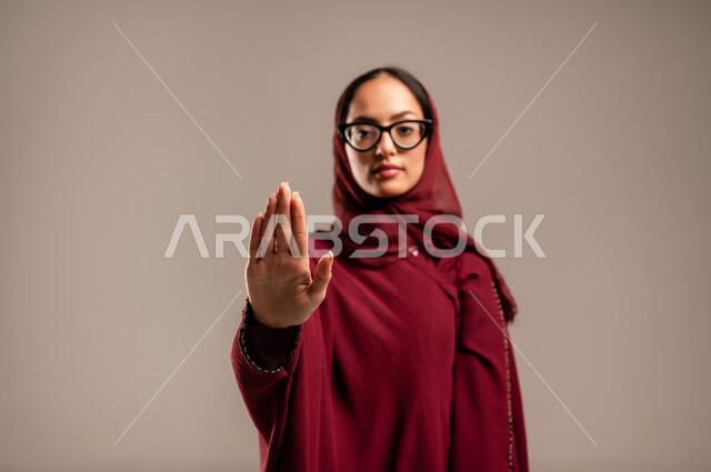 تعابير الرفض والإمتناع، الأمر بالتوقف عن فعل شيء ما، بورتريه مقرب لامرأه عربيه خليجيه سعوديه محجة ترتدي عباءة حمراء ترفع يدها بإيماءات تدل على النهي، الشعور بالإنزعاج والغضب، خلفية بيج