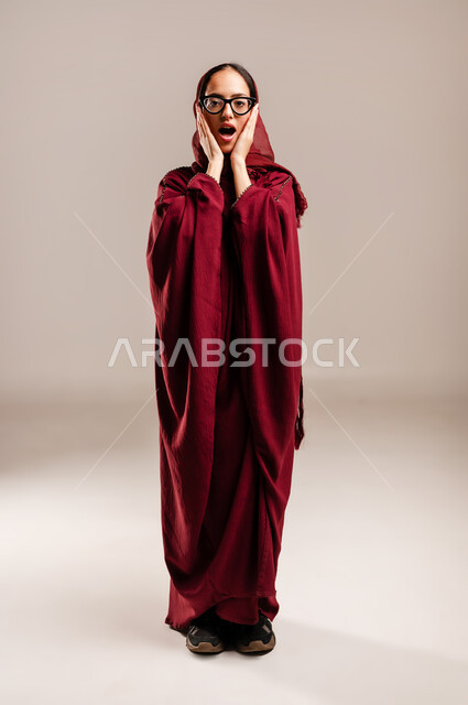 المفاجئه والذعر من شئ ما, ايماءات تدل على الذهول لسماع خبر مفاجئ، الصدمة والتعجب من حصول أمر ما، بورتريه لامرأة عربية خليجية سعودية محجبة ترتدي العبائه الحمراء تنظر إلى الكاميرا بتعابير الدهشة والخوف, الجسم بالطول الكامل، خلفية بيج