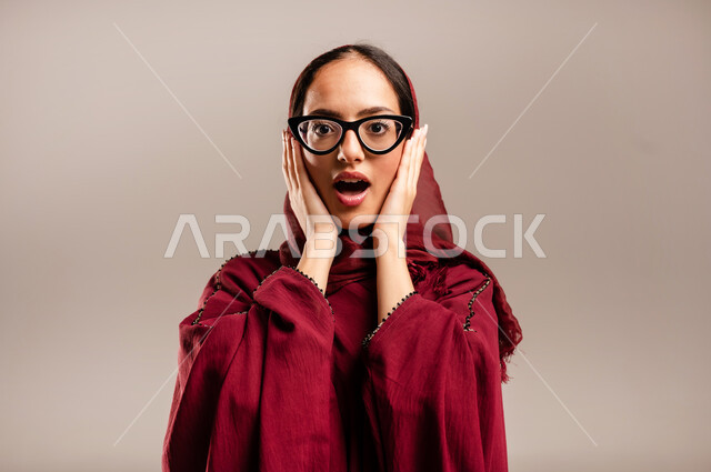المفاجئه والذعر من شئ ما, ايماءات تدل على الذهول لسماع خبر مفاجئ، الصدمة والتعجب من حصول أمر ما، صورة مقربة لامرأة عربية خليجية سعودية محجبة ترتدي العبائه الحمراء تنظر إلى الكاميرا بتعابير الدهشة والخوف, خلفيه بيج