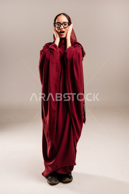 المفاجئه والذعر من شئ ما, ايماءات تدل على الذهول لسماع خبر مفاجئ، الصدمة والتعجب من حصول أمر ما، بورتريه لامرأة عربية خليجية سعودية محجبة ترتدي العبائه الحمراء تنظر إلى الكاميرا بتعابير الدهشة والخوف, الجسم بالطول الكامل، خلفية بيج
