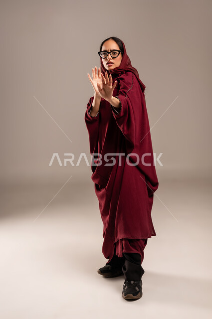 Command to stop doing something, expressions of refusal and abstention, portrait of a Saudi Arabian Gulf woman wearing a red abaya raising her hand with gestures indicating prohibition, feeling of annoyance and anger, full body, beige background