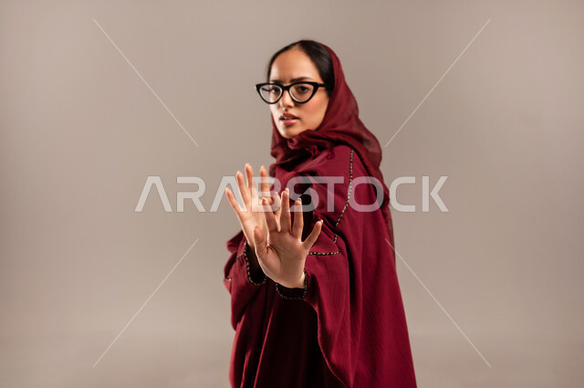 الأمر بالتوقف عن فعل شيء ما، تعابير الرفض والإمتناع، بورتريه مقرب لامرأه عربيه خليجيه سعوديه محجة ترتدي عباءة  حمراء ترفع يدها بإيماءات تدل على النهي، الشعور بالإنزعاج والغضب، خلفية بيج