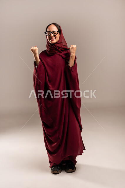 النجاح وتحقيق الأهداف، تعابير السرور والسعادة، بورتريه لامرأة عربية خليجية سعودية محجبة مبتسمة ترتدي العباءة الحمراء تنظر إلى الكاميرا بايماءات وجه ويدين تدل على الفوز والإنتصار، الجسم بالطول الكامل، خلفية بيج