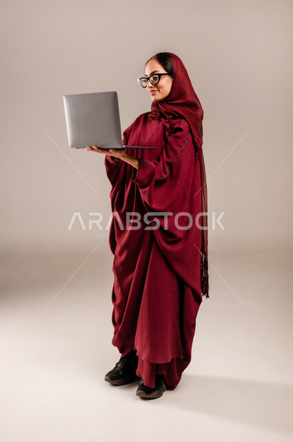 Using modern and advanced technical devices, the concept of remote work, a portrait of a veiled Saudi Arabian Gulf woman wearing a red abaya and glasses working on a laptop, completing tasks using modern technologies, full-length body, gray background