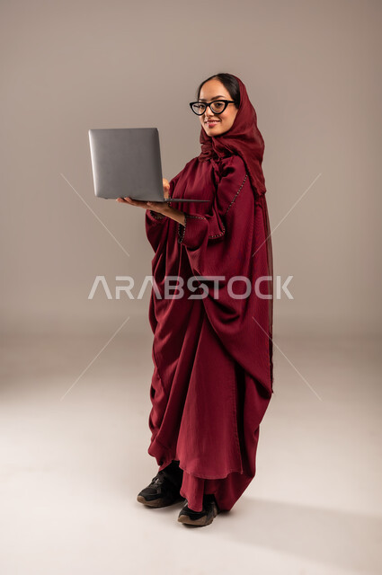 استخدام الأجهزة التقنية الحديثة والمتطورة, مفهوم العمل عن بعد, بورتريه لسيده عربية خليجية سعوديه محجبة ترتدي العباءة الحمراء والنظارات تعمل على جهاز الحاسوب المحمول, أنجاز المهام باستخدام التقنيات الحديثه, الجسم بالطول الكامل، خلفيه رمادية