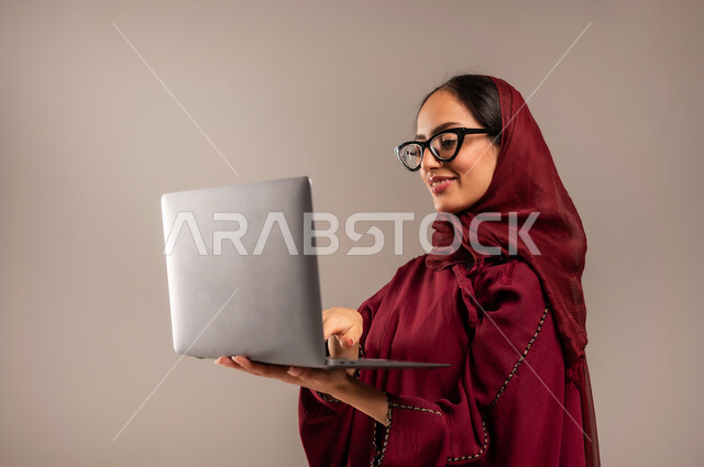 استخدام الأجهزة التقنية الحديثة والمتطورة, مفهوم العمل عن بعد, بورتريه مقرب لسيده عربية خليجية سعوديه محجبة ترتدي العباءة الحمراء تعمل على جهاز الحاسوب المحمول, أنجاز المهام باستخدام التقنيات الحديثه, خلفيه بيج