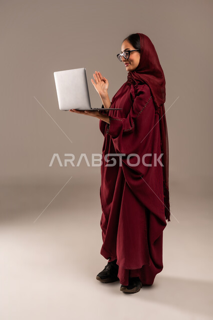 استخدام الأجهزة التقنية الحديثة والمتطورة, مفهوم العمل عن بعد, بورتريه لسيده عربية خليجية سعوديه محجبة ترتدي العباءة الحمراء والنظارات تعمل على جهاز الحاسوب المحمول, أنجاز المهام باستخدام التقنيات الحديثه, الجسم بالطول الكامل، خلفيه رمادية