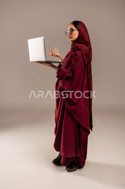 استخدام الأجهزة التقنية الحديثة والمتطورة, مفهوم العمل عن بعد, بورتريه لسيده عربية خليجية سعوديه محجبة ترتدي العباءة الحمراء والنظارات تعمل على جهاز الحاسوب المحمول, أنجاز المهام باستخدام التقنيات الحديثه, الجسم بالطول الكامل، خلفيه رمادية