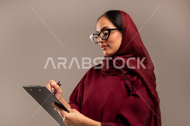 مفهوم كتابه وتدوين الملاحظات، بورتريه مقرب لامرأة عربية خليجية سعودية ترتدي الحجاب والعباءة الحمراء تمسك بيدها قلم تقوم بالكتابة على نوت تنظر إلى الكاميرابايماءات التفكير والتأمل، خلفيه بيج