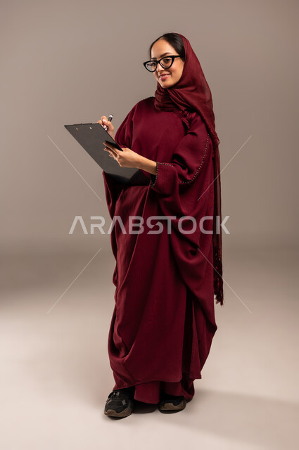 Concept of writing and taking notes, portrait of a Saudi Arabian Gulf woman wearing hijab and red abaya holding a pen in her hand writing on a notepad looking at the camera with gestures of thinking and contemplation, beige background