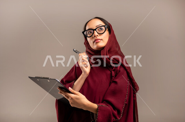 مفهوم كتابه وتدوين الملاحظات، بورتريه مقرب لامرأة عربية خليجية سعودية ترتدي الحجاب والعباءة الحمراء تمسك بيدها قلم تقوم بالكتابة على نوت تنظر إلى شيء ما بايماءات التفكير والتأمل، خلفيه بيج