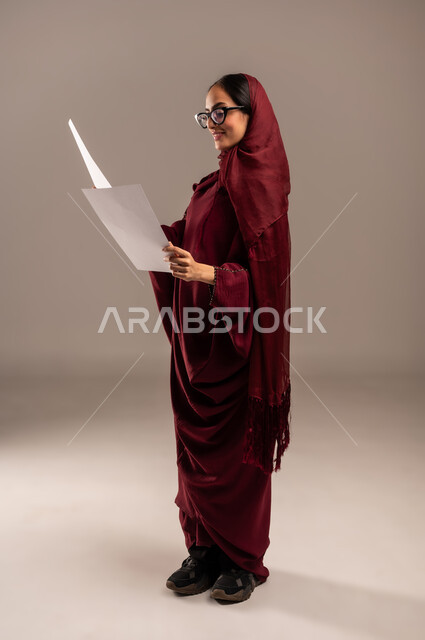 اعداد التقارير الخاصة بالعمل، بورتريه من الجانب لامرأة عربية خليجية سعودية محجبة ترتدي العباءة الحمراء تحمل بيدها مجموعة من الورق الأبيض تنظر اليها، مراجعة ومتابعة الخطط والمشاريع، تبادل الخبرات والمهارات، الجسم بالطول الكامل، خلفية بيج