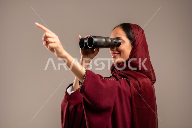 رؤية الاشياء البعيدة عبر التلسكوب، التخطيط المستقبلي للأهداف و الطموحات، حب الاستكشاف والمغامرة، بورتريه مقرب لامرأة عربية خليجية سعودية محجبة ترتدي العباءة الحمراء تنظر من خلال منظار ثنائي العينين, خلفيه بيج