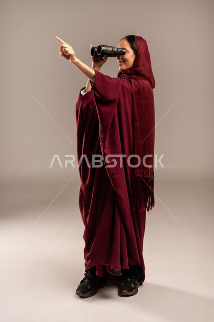 Seeing distant objects through a telescope, future planning for goals and ambitions, love of exploration and adventure, portrait of a veiled Saudi Arabian Gulf woman wearing a red abaya looking through binoculars, full body, beige background